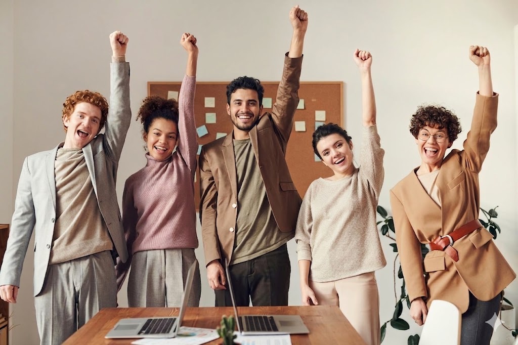 Equipo diverso de cinco personas celebrando un logro compartido en oficina, con gesto de victoria en un ambiente corporativo luminoso.