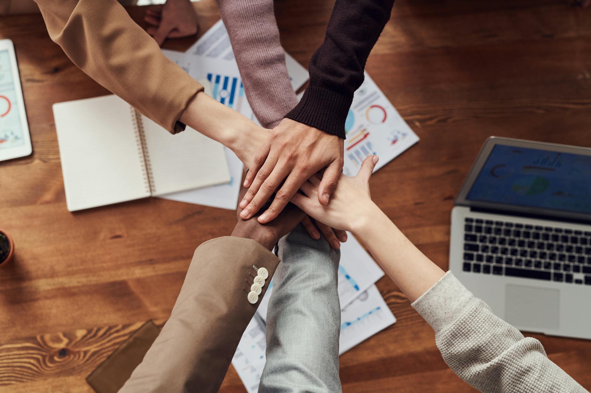 Manos de un equipo unidas en señal de compromiso y colaboración, sobre una mesa de trabajo con materiales de planificación.